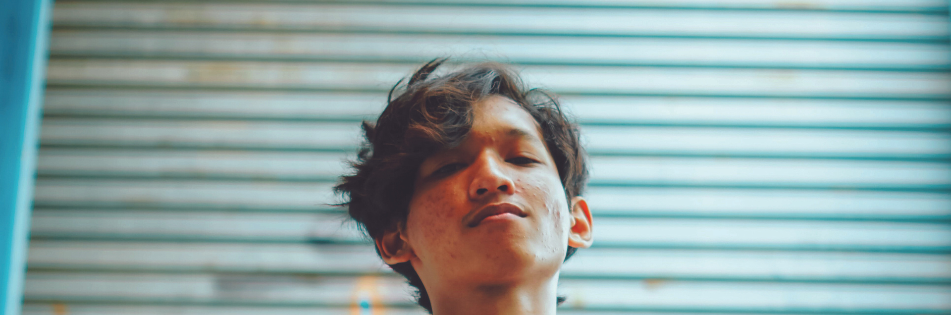 Une jeune personne à l'extérieur fait face à la caméra, avec en arrière-plan un mur métallique recouvert de graffitis.